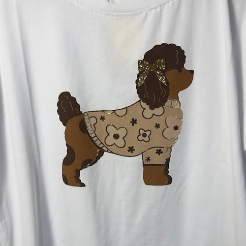 Camiseta blanca de PINTONAS con un caniche marrón con un suéter estampado de flores y un lazo brillante en la cabeza.